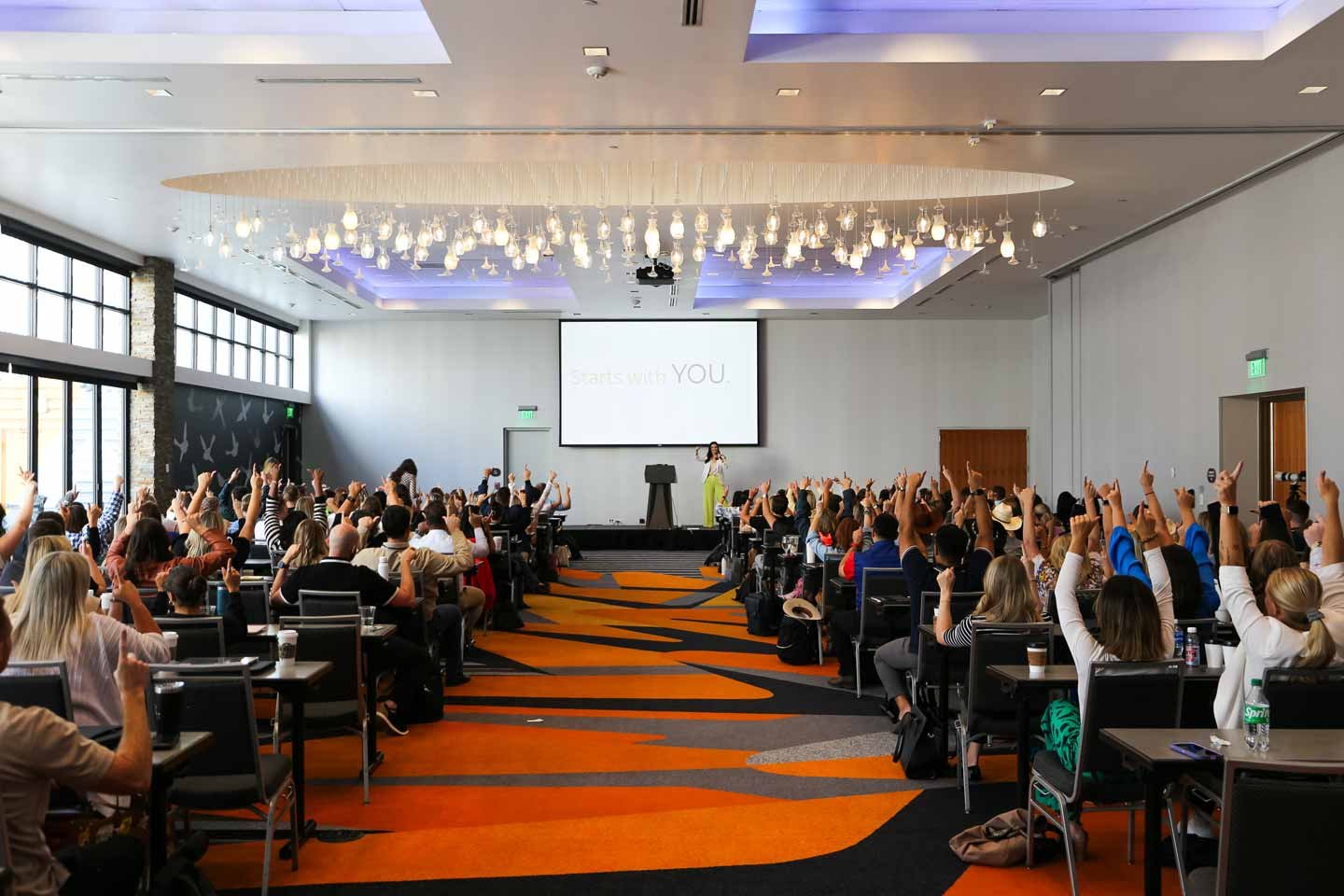 Amy Reczek Inspiring a Full Audience Amy Reczek speaking on stage to a large audience with raised hands during an interactive session.