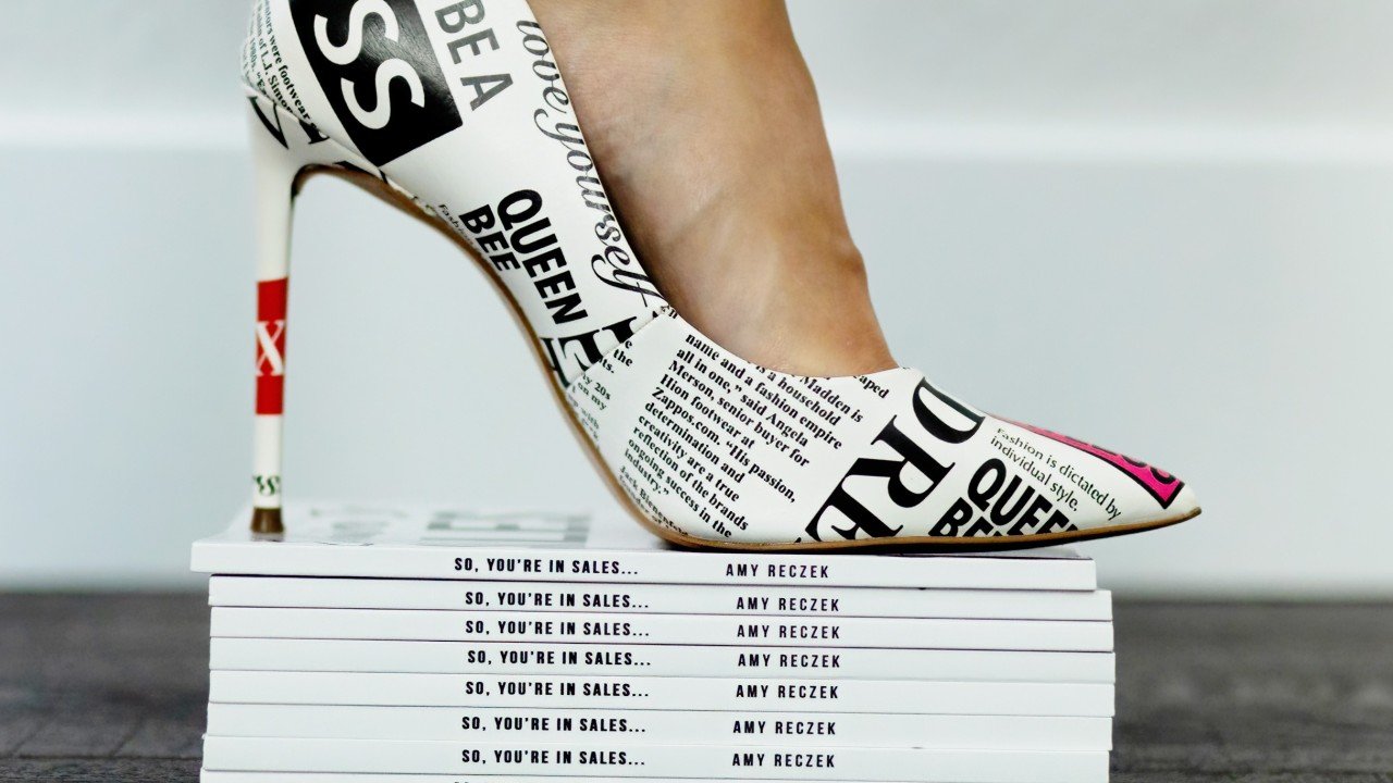 Close-up of a custom high heel with bold typography resting on a stack of Amy Reczek’s book So, You’re in Sales...